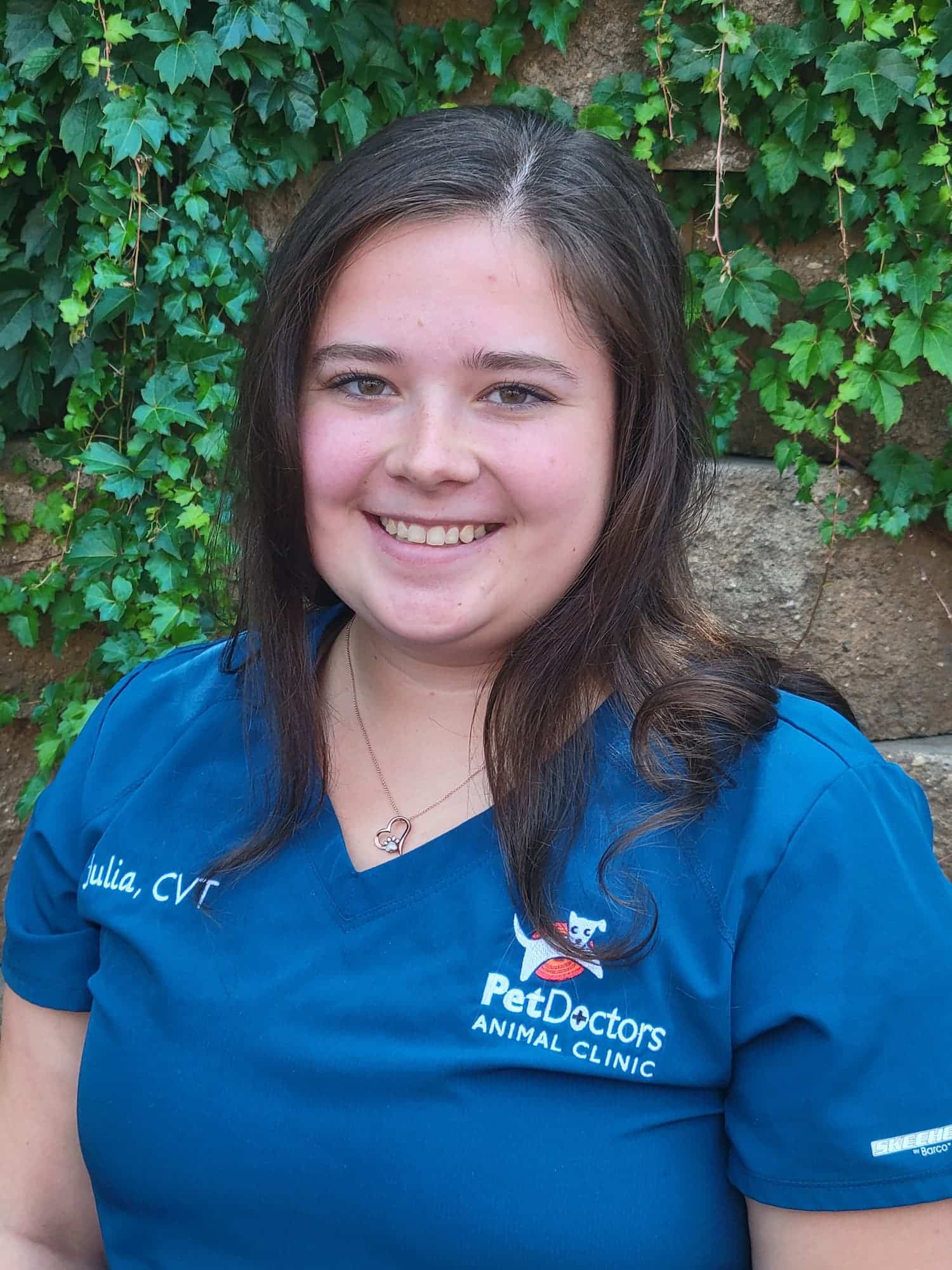 A woman wearing a blue "Pet Doctors Animal Clinic" uniform stands in front of a stone wall with green ivy.