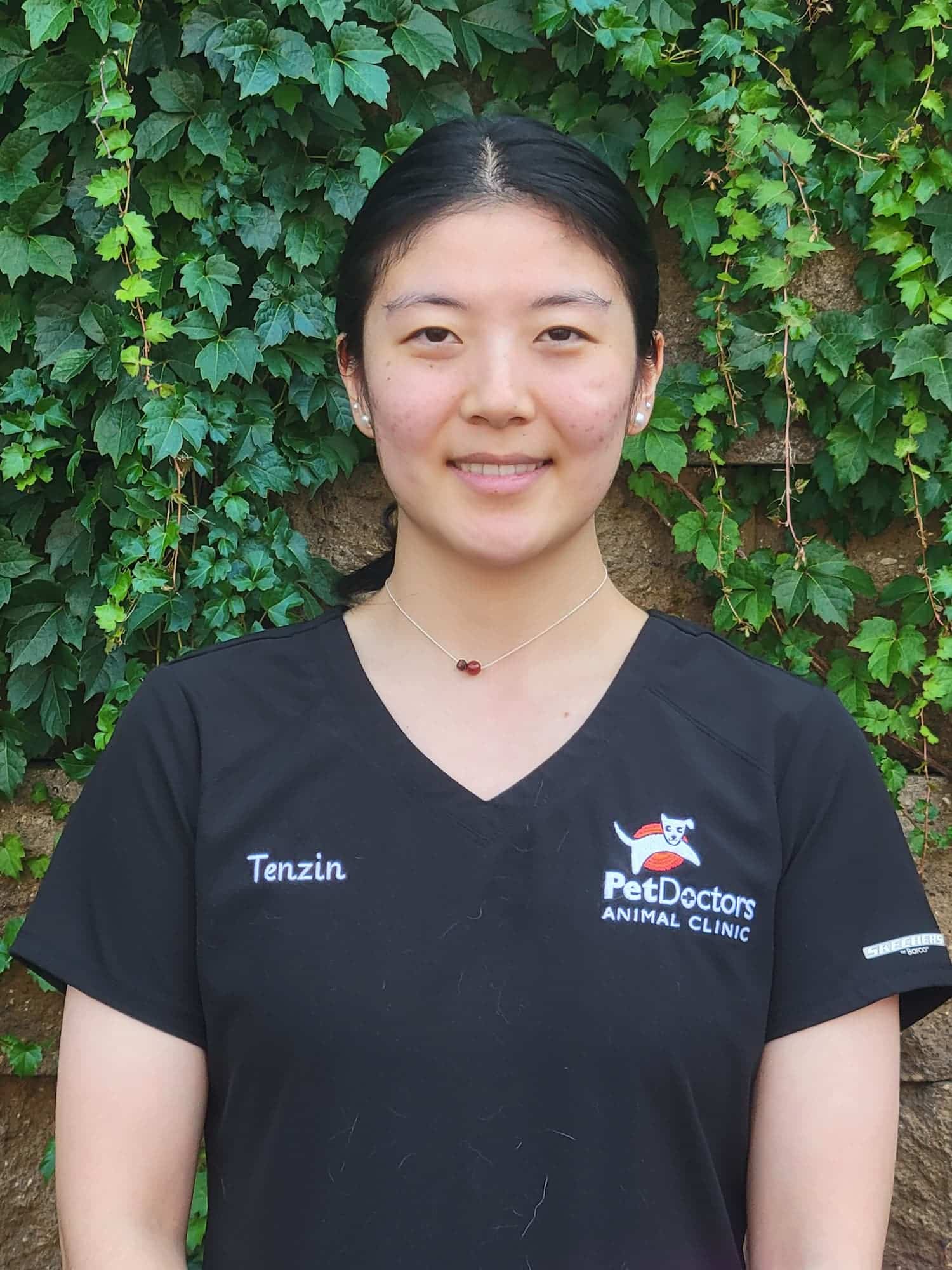 A woman wearing a black "Pet Doctors Animal Clinic" uniform with the name "Tenzin" stands in front of a wall covered with green ivy.
