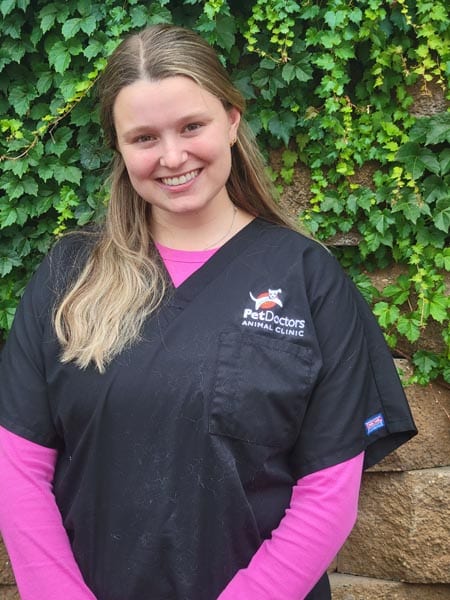 A woman wearing a black "Pet Doctors Animal Clinic" scrub top and pink undershirt stands in front of a wall covered with green ivy.