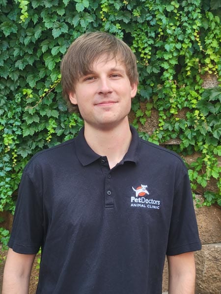 A person wearing a black "PetDoctors Animal Clinic" polo shirt stands in front of a wall covered with green ivy.
