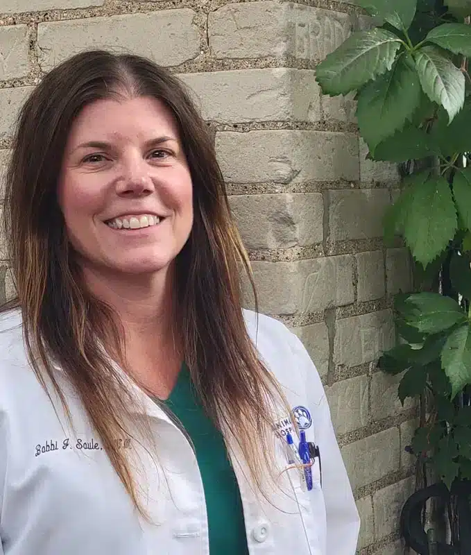 A woman with long brown hair wearing a white lab coat stands in front of a brick wall with green plants nearby, smiling at the camera.