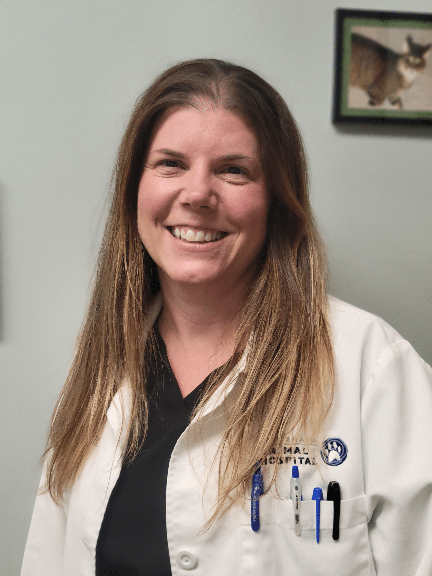 A person with long hair, wearing a white coat with pens in the pocket, smiles at the camera. A small framed picture of a cat is on the wall behind them.