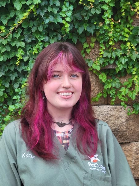 A person with pink-highlighted hair wearing a green PetDocs Animal Clinic uniform stands in front of a leafy wall. The name "Katie" is embroidered on the shirt.