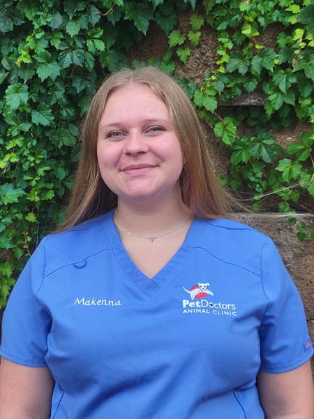 A person wearing a blue Pet Doctors Animal Clinic uniform top with the name "Makenna" stands in front of a leafy green background.