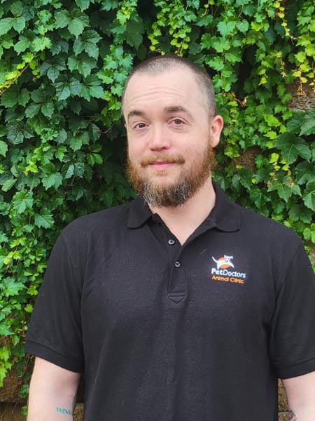 A person with a beard wearing a black "Pet Doctors Animal Care" polo shirt stands in front of a wall covered with green ivy leaves.