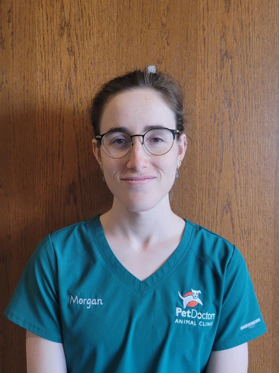 Person with glasses and brown hair, wearing a teal shirt with "Pet Doctors Animal Clinic" logo and "Morgan" embroidered, stands against a wooden background.