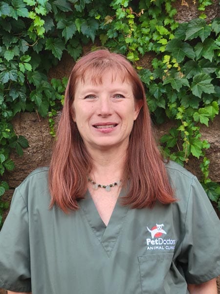 A woman with straight red hair, wearing green scrubs with a "Pet Doctors Animal Clinic" logo, stands in front of a leafy background.