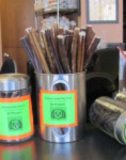 Metal container with assorted sticks on a countertop, surrounded by jars with bright labels.