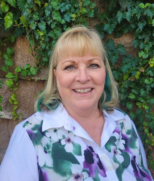A woman with blonde hair, wearing a white shirt with a purple and green floral pattern, smiles in front of a wall covered with green ivy.