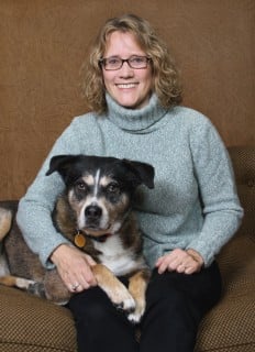 A person with glasses and curly hair smiles while sitting on a couch with a large dog resting its head on their lap.