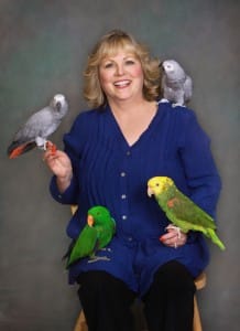 A woman in a blue shirt sits with four parrots: two gray on her shoulders and two green on her lap.