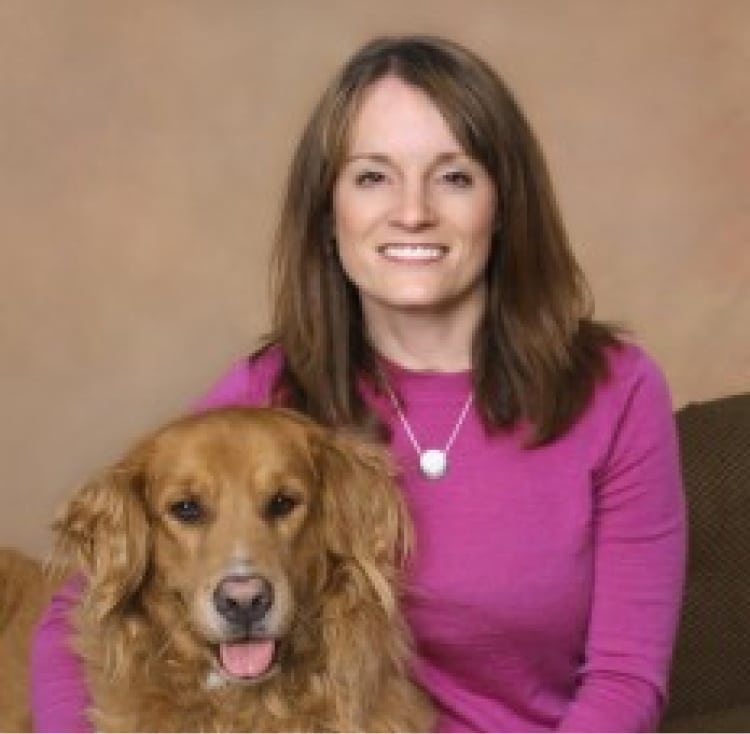 A woman in a pink sweater smiles while sitting next to a golden retriever against a beige background.