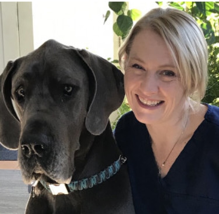 A woman with blonde hair smiles next to a large gray Great Dane indoors.