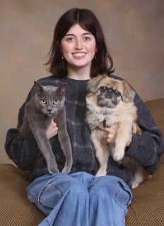 A person sitting on a couch holding a gray cat and a small fluffy dog, posing for a portrait.