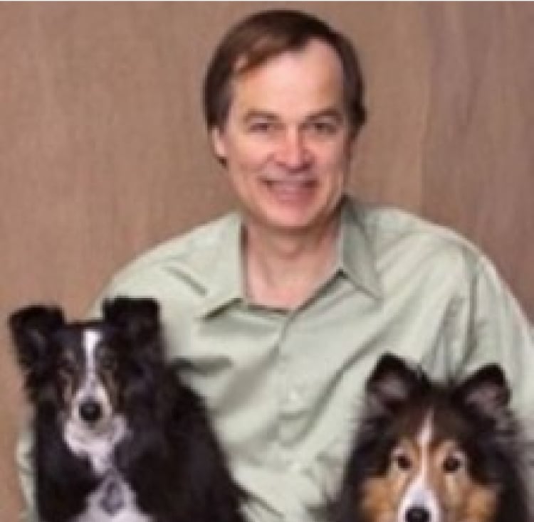 Man in a light green shirt smiling, seated with two fluffy dogs.